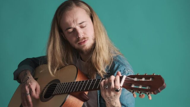 Long haired blond male musician looking sensual playing on acoustic guitar and singing over colorful background. Music concept. Creative guy
