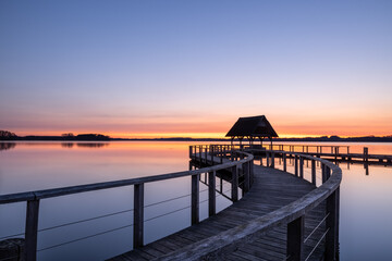 Curvy Pier construction and shelter with thatched roof at beautiful colorful sunrise under clear sky at Lake Hemmelsdorf, Schleswig-Holstein, Northern Germany