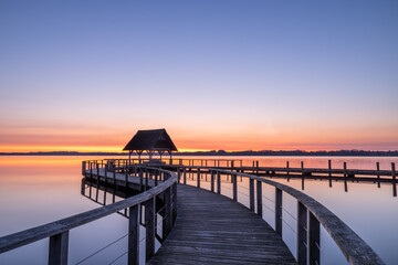 Swinging curved Pier construction and shelter with thatched roof at beautiful red dawn under clear sky at Lake Hemmelsdorf, Schleswig-Holstein, Northern Germany