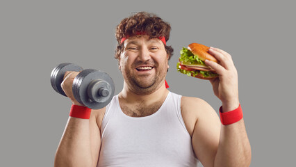 Funny smiling man with dumbbell and delicious burger looking at camera. Happy fat guy holding free...