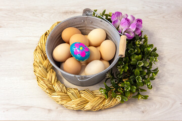 Metal bucket with chicken eggs and a colorful, hand-painted Easter egg. Easter composition with flowers and a straw wreath on a wooden background.