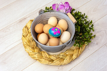 Metal bucket with chicken eggs and a colorful, hand-painted Easter egg. Easter composition with flowers and straw wreath on a wooden background.