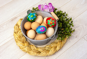 A beautiful Easter, decorative composition with a metal bucket filled with chicken eggs and Easter eggs. Hand-painted colorful Easter eggs. Decorative buckets outdoors with flowers on a wooden backgro