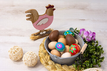 Colorful Easter, decorative composition on a wooden background. Metal bucket filled with chicken eggs, Easter eggs and chocolate eggs. In the background, a hand-painted wooden hen.