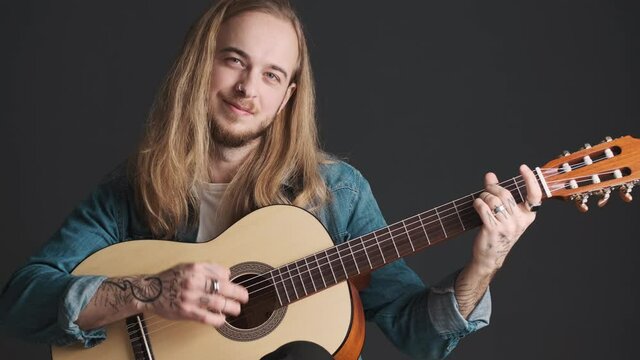Young long haired blond musician playing on acoustic guitar looking happy over black background. Cool guy working in studio 