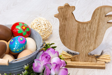 A wooden hen is looking at an Easter colorful decoration with a metal bucket, colorful Easter eggs and a chocolate egg. All surrounded by flowers on a wooden background.