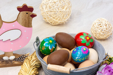 A wooden hen is looking at an Easter colorful decoration with a metal bucket, colorful Easter eggs and a chocolate egg. All surrounded by flowers on a wooden background.