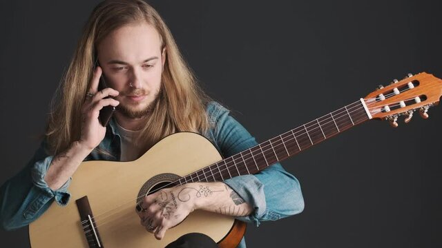 Young bearded male musician with guitar talking on smartphone during rehearsal in studio isolated on black background. Music concept