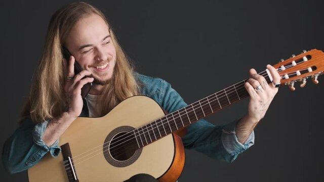 Young long haired male musician talking on smartphone while tuning the guitar over black background. Music concept