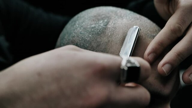 Professional barber shaves customer head with straight razor. Beard cut with old-fashioned blade at barber shop.