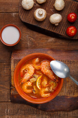 Tom yam soup with shrimps in a plate on a wooden table next to a bowl with coconut milk mushrooms and tomatoes.