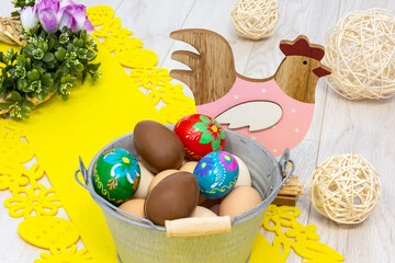 Easter composition full of colorful elements. Among them, a colorful hand-painted hen, a metal bucket filled with chicken eggs and colorful Easter eggs. Everything on a light wooden floor.