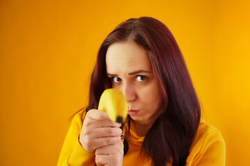 Portrait of young woman with banana on yellow background. Close up of female in yellow hoodie plays with fruit, imagining it as weapon.