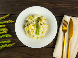 Risotto with asparagus. Dish with creamy rice with asparagus. Mediterranean food. Plate with cutlery, fork and knife. Spring recipe. Rustic wooden table. Flat lay. Top view. 