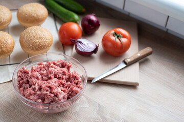 Ground beef in a glass bowl on the kitchen table with other hamburger ingredients, cooking at home.