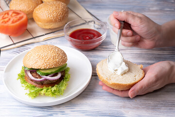 Female hands smearing sauce on bun on white wood background with beef burger, cooking hamburger at home.