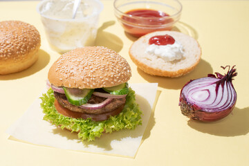 Delicious hamburger on a on a yellow background with ingredients for a burger: ketchup, sauce, vegetables.