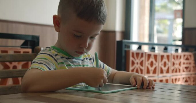 A portrait of a child sits at a table at home and draws on his tablet with a special pen, a preschooler uses a special tablet for drawing.