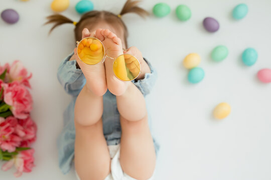 A child lies on a white background with Easter colored eggs and plays with yellow glasses. The girl with the ponytails put her glasses on her feet. The baby hides her face.