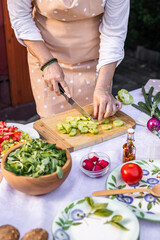 Female chef with apron cutting cucumber for vegetable salad. Woman cooking healthy mediterranean food outdoors