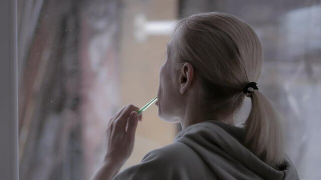 A woman brushes her teeth while holding a toothbrush in her left hand, standing by the window