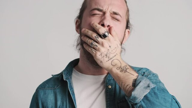 Portrait of attractive bearded guy with tattooed hand yawning on camera can't wake up in the morning over white background. Sleepy man