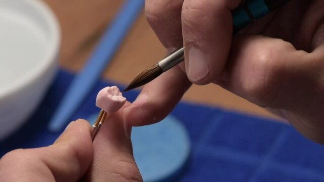 Dental technician applies ceramic material to the crown of a dental implant in a dental laboratory using tweezers and a brush. Dental technology close-up.