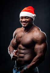 Happy sexy Santa Claus in red hat posing on black background. Muscular bodybuilder with naked torso looking at camera.