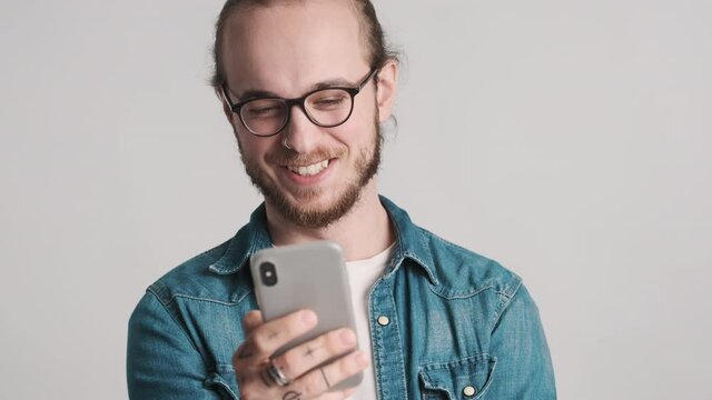 Portrait of positive bearded guy in eyeglasses dressed in casual wear looking happy reading latest news on smartphone over white background