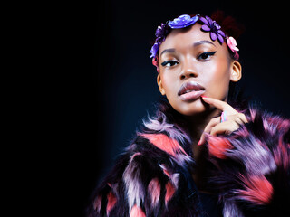 young pretty african american woman in spotted fur coat and flowers on head smiling on black background, fashion people concept