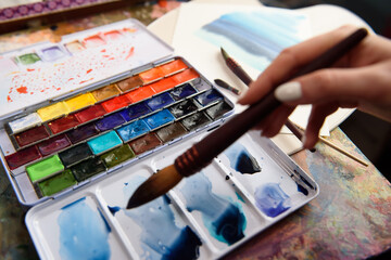 Fototapeta premium Set of bright saturated watercolor paints and brushes for painting. The artist mixes different shades of paints on a plastic palette. Drawing and creative tools close-up. Top view, selective focus.