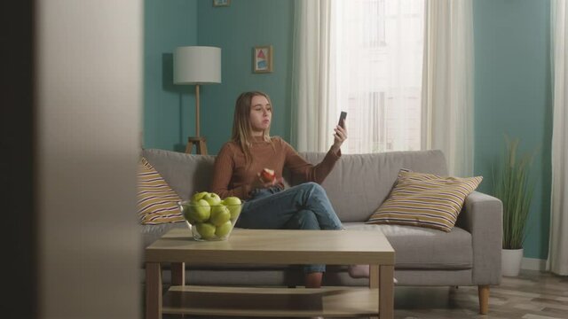 Young girl in blue jeans and brown sweater sits on beige sofa on window background, bites red apple and talks on video call. On coffee table is glass jar with green apples. Camera dollies horizontally