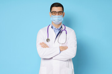 Portrait of young male doctor wearing medical mask, standing with crossed arms in white coat, isolated on blue background