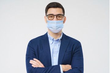 Young business man wearing glasses and medical mask, standing with crossed arms isolated on gray background