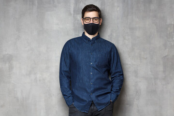 Young man wearing denim shirt, glasses and black mask, standing against gray textured wall