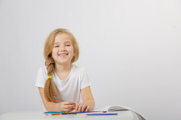 A small cute schoolgirl girl with blonde hair draws an inscription in an album with colored pencils on a light background
