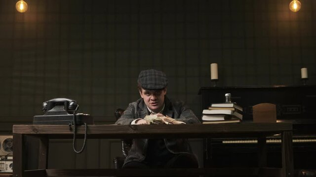 gangster or bandit sits at table in room with green walls, gets up and shouts at his interlocutor. man in plaid jacket and classic cap screaming and very angry. concept of early 20th century.