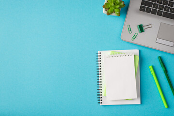 Top view photo of workplace with laptop green chancellery notebooks plant on isolated blue background with copyspace