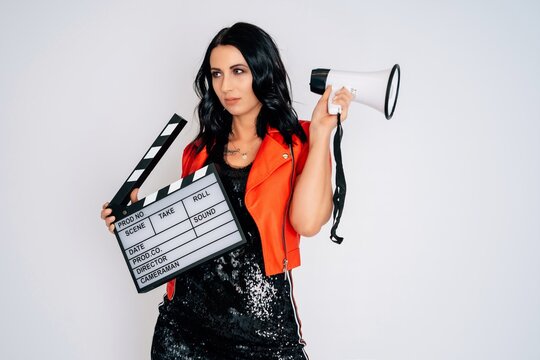 young woman in black dress with red jacket and high heels holding clapperboard screaming in megaphone isolated on white background