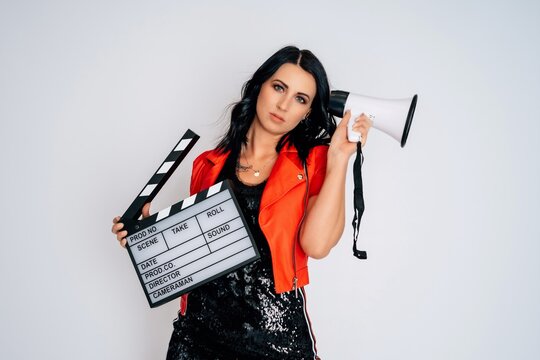 young woman in black dress with red jacket and high heels holding clapperboard screaming in megaphone isolated on white background