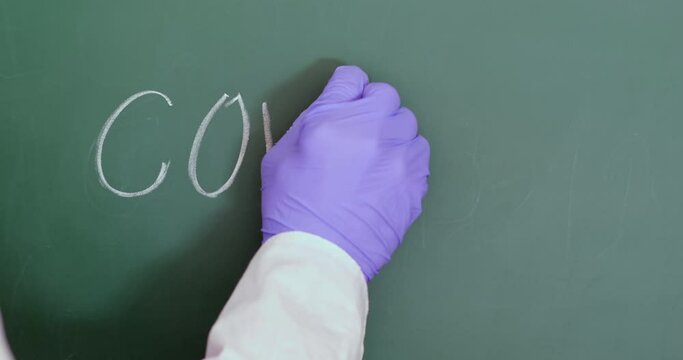 Close-up, man in a robe and blue gloves writes "COVID-19" on a blackboard in chalk and underlines it.