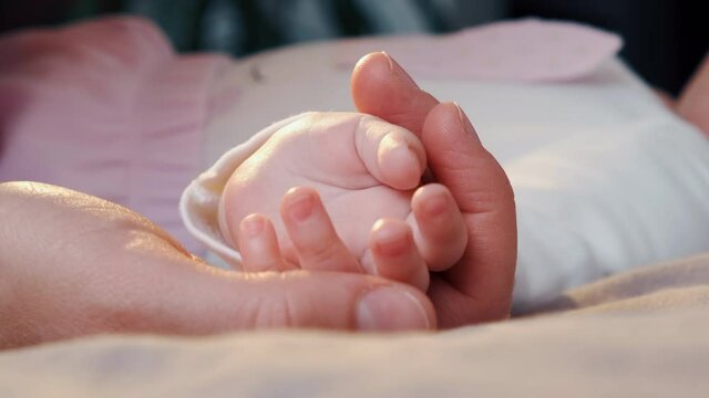 Close up of caring young mother holds and touches baby hand lying on cozy bed at home. Happy loving mum and funny adorable infant child girl relaxing in bedroom. Motherhood and childcare concept