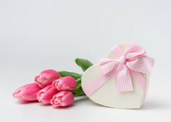 Fototapeta na wymiar Beautiful tulips of pink color in a silk ribbon near a heart-shaped gift box with a ribbon and a beautifully tied pink bow. The concept of a gift and surprise for a loved one.
