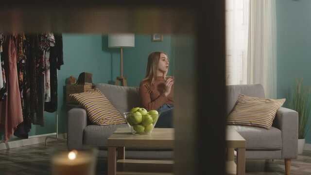 Young girl in blue jeans and brown sweater sits on beige sofa on window background, bites red apple and talks on video call. On coffee table is glass jar with green apples. Camera dollies horizontally