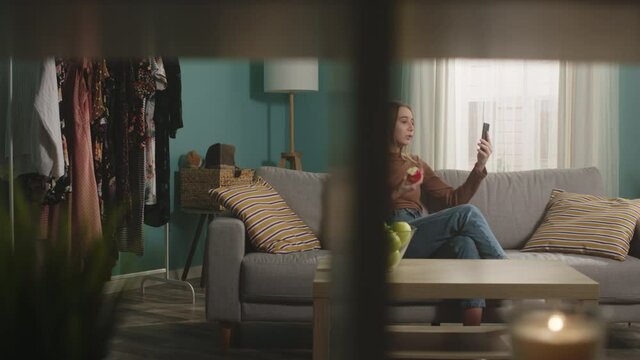 Young girl in blue jeans and brown sweater sits on beige sofa on window background, bites red apple and talks on video call. On coffee table is glass jar with green apples. Camera dollies horizontally