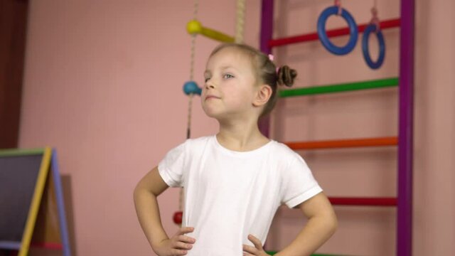 Little beautiful girl in a white T-shirt performs head rotation physical exercise.Exercise for the muscles of the neck and back.Sports at home.Girl in the gym.Morning exercises for children.