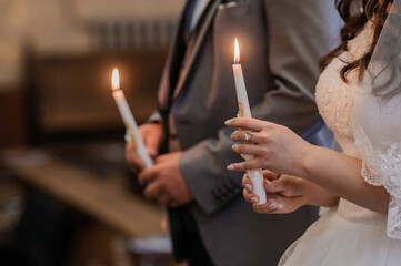 The bride and groom hold shining candles during the ceremony in the church. Hands of newlyweds with candles in the church. Church religious details. Traditions