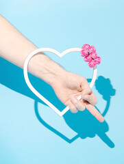 Hand holding white heart frame decorated with pink roses, showing middle finger. Pastel blue background. Minimal creative idea.