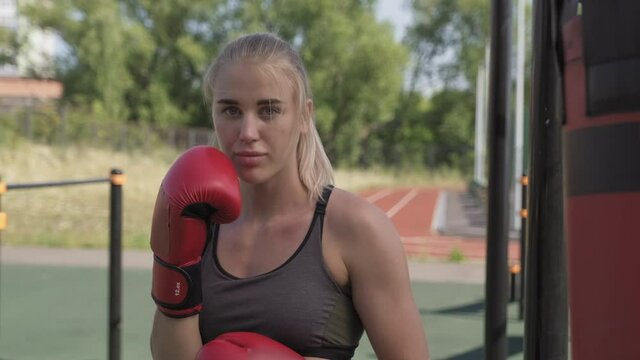 Medium shot of concentrated female boxer boxing punch bag outdoors then looking at camera