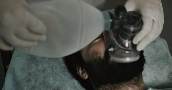 Close up of medics hands putting on ill bearded man oxygen inhalation mask. Top view of unconscious guy in 30s mumbling while lying on medical transportation cart on way to hospital.